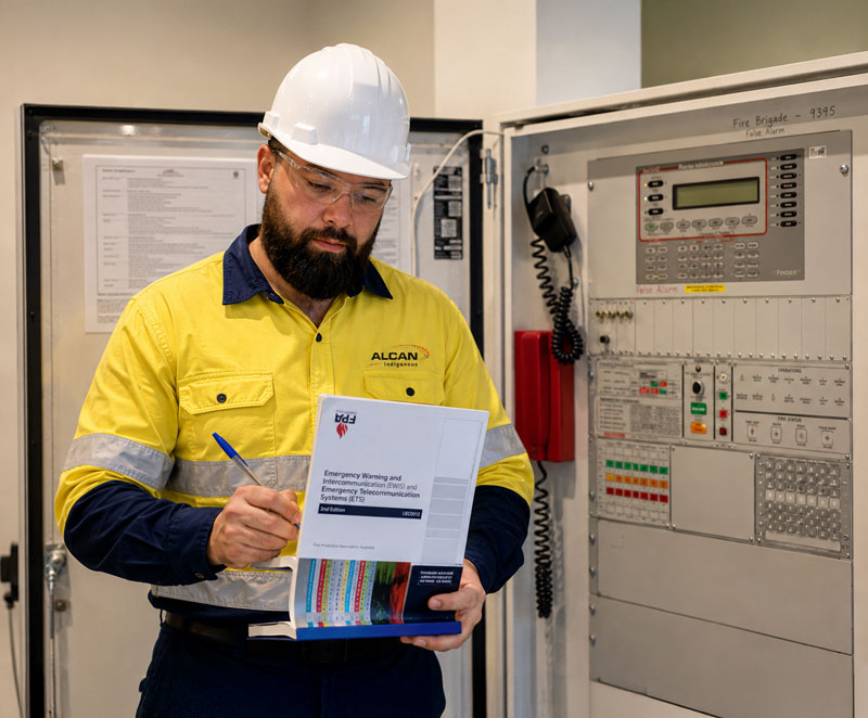 Terrence, a fire safety technician in high-visibility workwear and safety helmet, reviewing documentation while inspecting an EWIS fire alarm control panel.